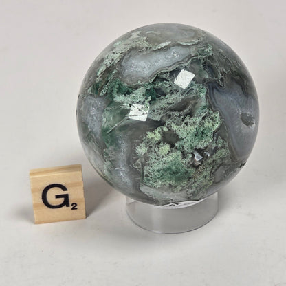 A polished round stone sphere with green and gray marbled patterns, displayed on a clear stand next to a small wooden letter tile spelling G2, on a light gray surface.