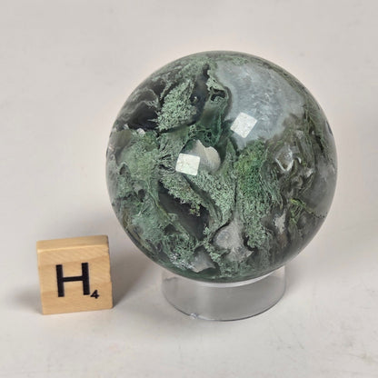 A polished green stone sphere with natural marbled patterns, displayed on a clear acrylic stand next to a small wooden letter block with the letter H, set against a light gray surface.