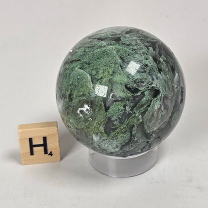 A polished green stone sphere with natural marbled veining, displayed on a clear acrylic stand next to a small wooden letter block with the letter H, on a light gray surface.