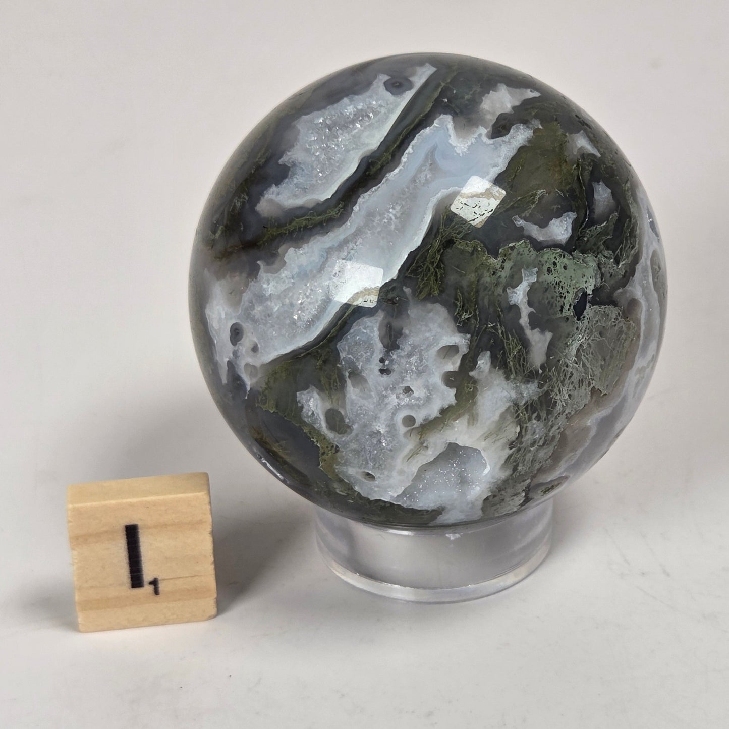 A polished agate crystal sphere with natural marbled patterns of gray, white, and olive green, displayed on a clear acrylic stand next to a small wooden block measuring marker on a light gray surface.