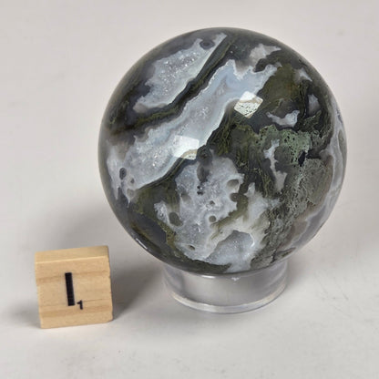 A polished agate crystal sphere with natural marbled patterns of gray, white, and olive green, displayed on a clear acrylic stand next to a small wooden block measuring marker on a light gray surface.