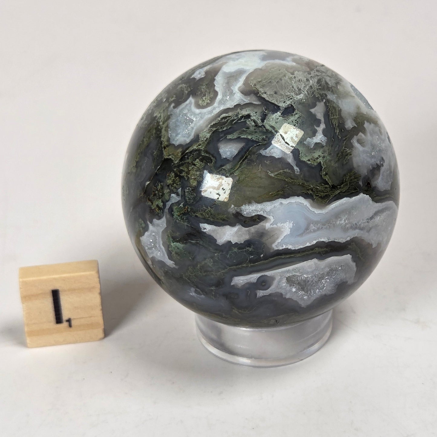 A polished geode sphere with dark gray, green, and white marbled patterns, displayed on a clear stand next to a small wooden block for scale, on a light gray background.