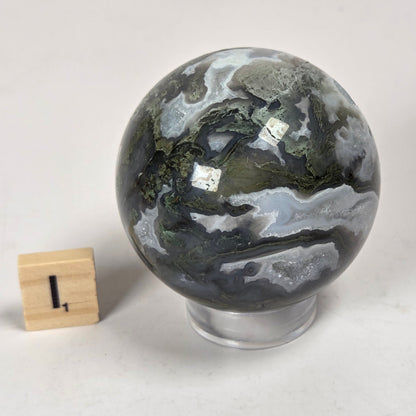 A polished geode sphere with dark gray, green, and white marbled patterns, displayed on a clear stand next to a small wooden block for scale, on a light gray background.