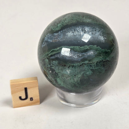 A polished dark green and black agate crystal sphere on a clear acrylic stand, shown next to a wooden Scrabble tile with the letter J on a light gray background.
