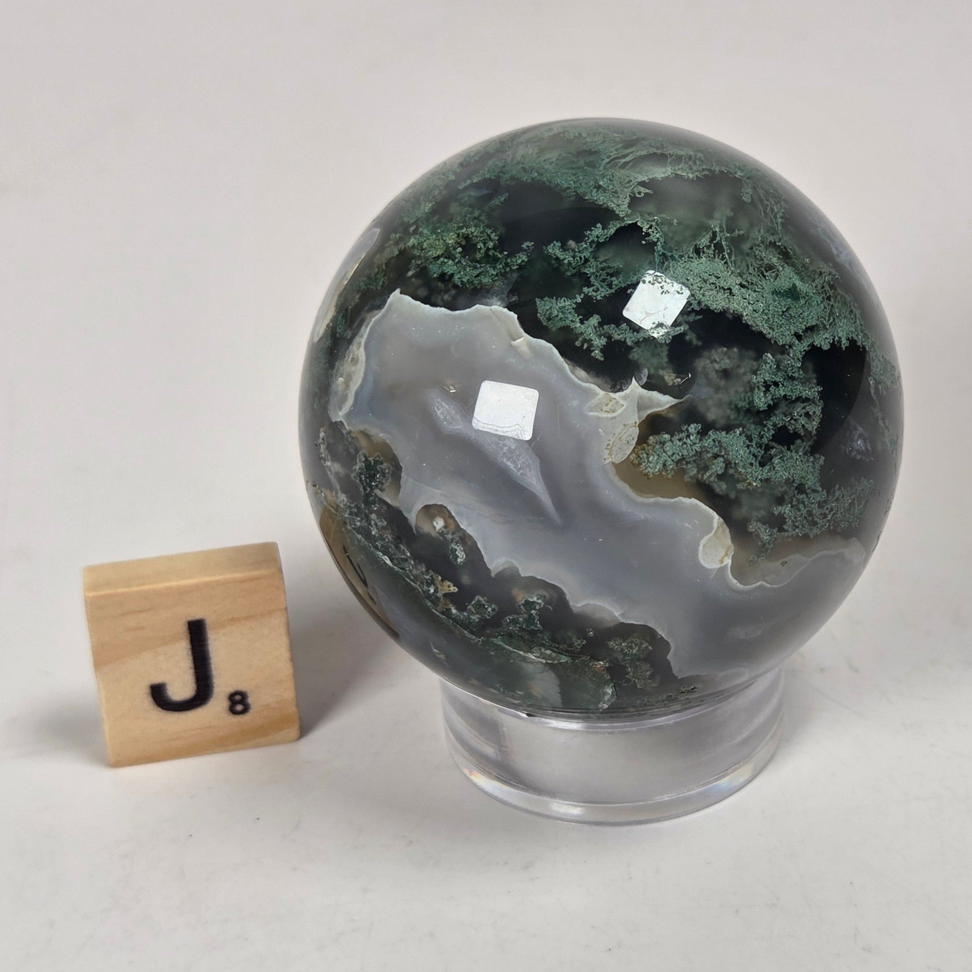 A polished agate crystal sphere with green and white banding, displayed on a clear acrylic stand next to a small wooden scrabble tile on a light gray background.