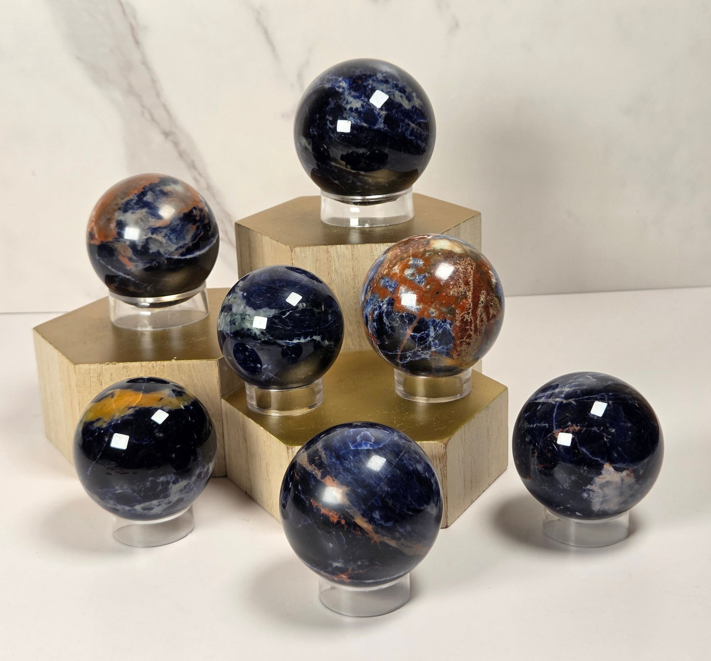 Seven polished crystal spheres in various colors, including deep blue, brown, and orange, displayed on clear stands atop gold-colored geometric pedestals against a white marble background.
