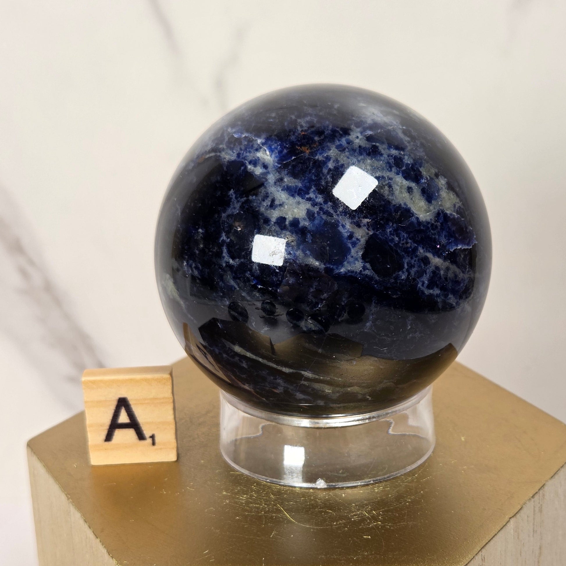 A polished dark blue lapis lazuli crystal sphere on a clear acrylic stand, displayed on a gold hexagonal pedestal next to a small wooden letter tile.