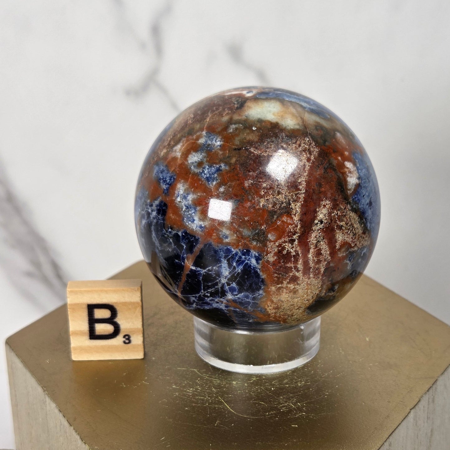 A polished natural stone sphere with swirling brown, gold, and deep blue marbling sits on a clear acrylic stand atop a light wood pedestal. A wooden Scrabble tile with the letter B is placed beside it against a light marble-patterned background.