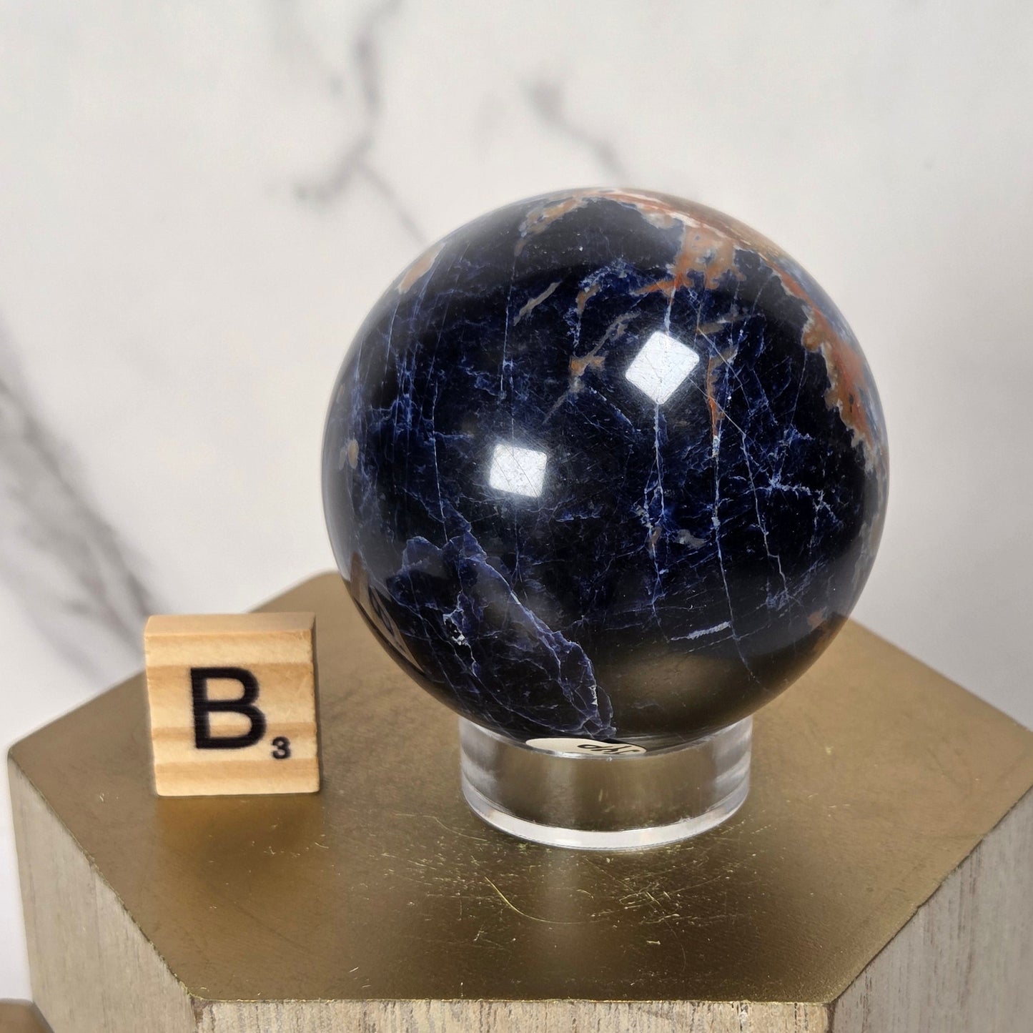 A polished dark blue and black stone sphere with natural veining, displayed on a clear acrylic stand atop a gold-toned geometric pedestal. A small wooden Scrabble tile with the letter B is placed beside it against a light marble-patterned background.