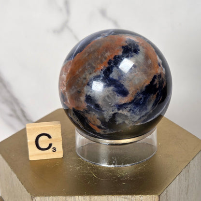 A polished round stone sphere with marbled dark blue, brown, and gray patterns sits on a clear acrylic stand atop a gold hexagonal display block. A small Scrabble tile with the letter C3 rests beside the stand against a light marble-patterned background.