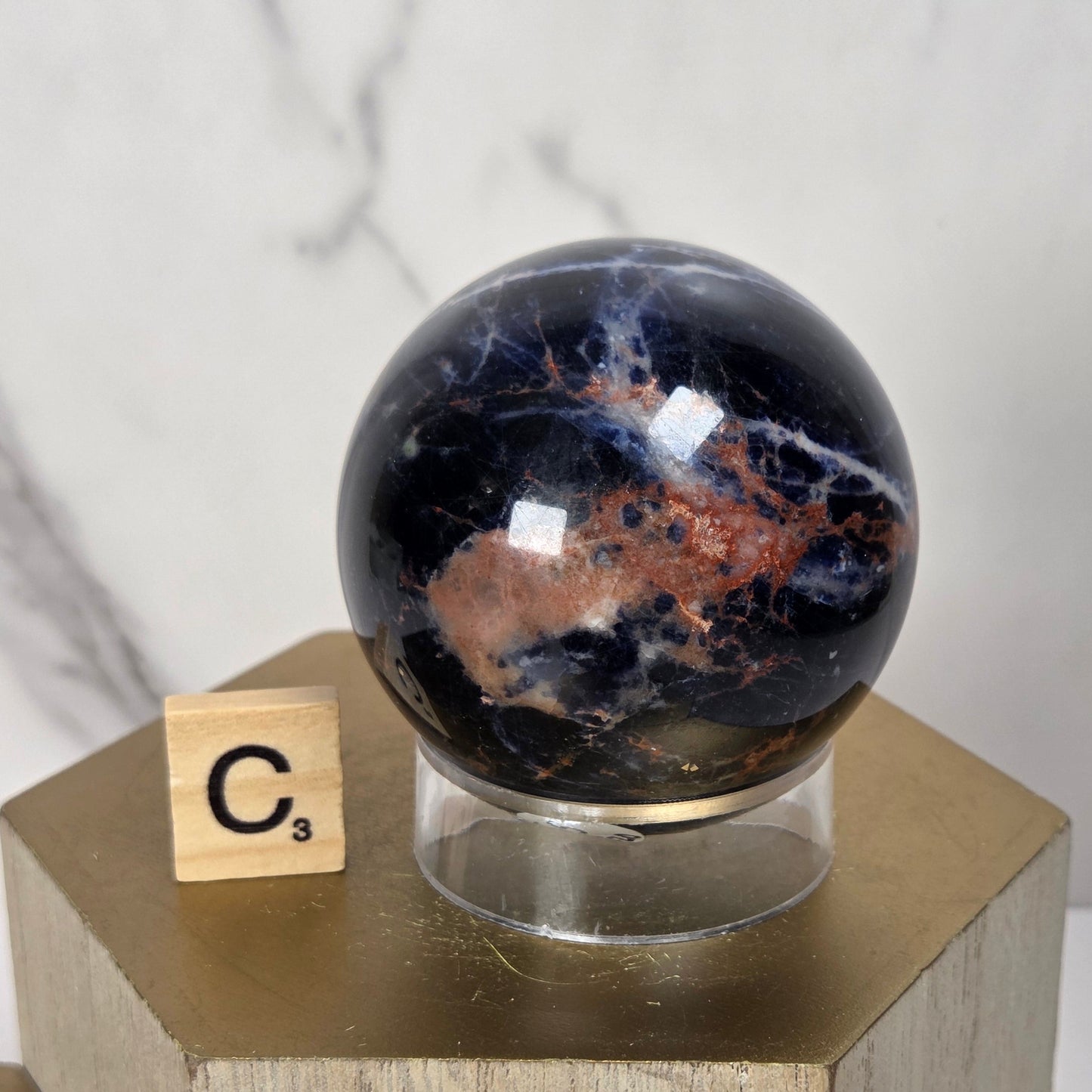 A polished dark stone sphere with coppery orange and white veining, displayed on a clear acrylic stand atop a gold-toned geometric pedestal. A small Scrabble tile showing 'C3' sits beside it against a light marble-patterned background.