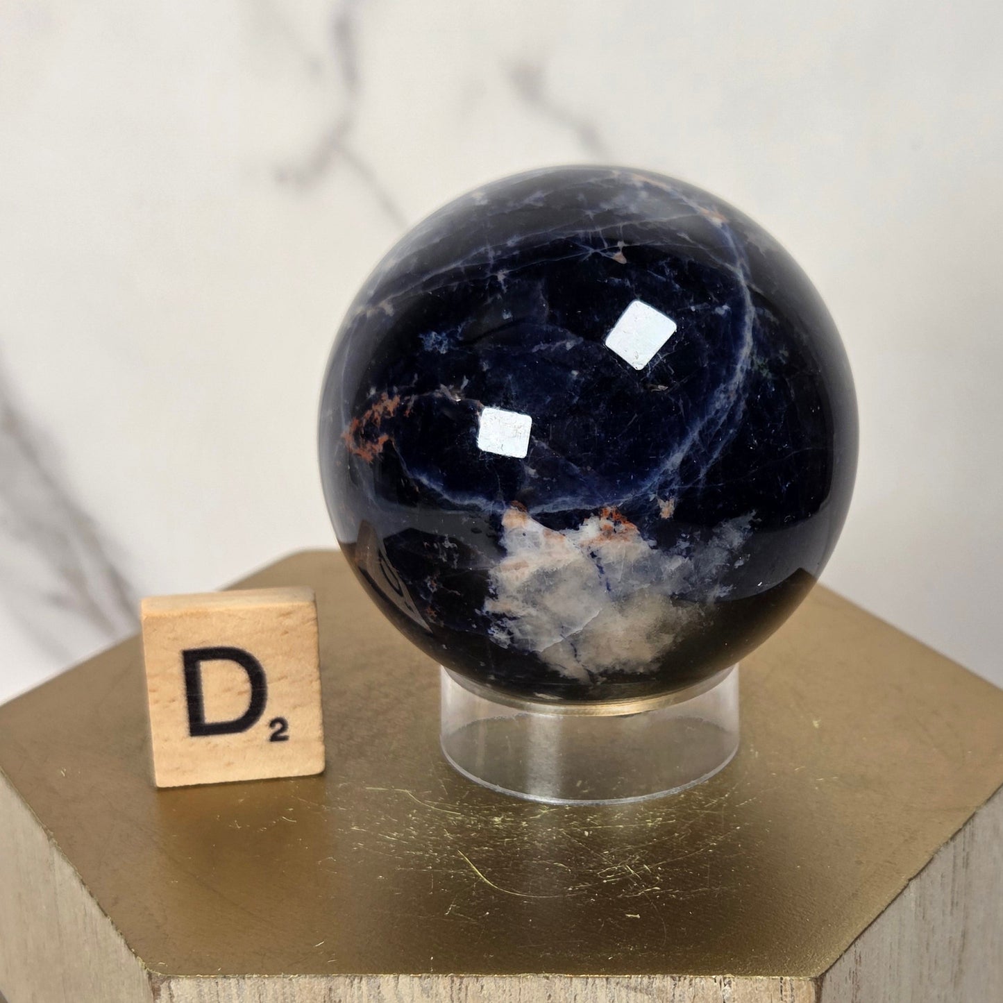 A polished dark blue jasper sphere on a clear acrylic stand, displayed on a gold-colored geometric pedestal next to a small Scrabble tile showing the letter D2, against a light marble background.
