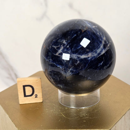 A polished dark blue sapphire crystal sphere displayed on a clear acrylic stand atop a gold-colored geometric pedestal, with a small Scrabble tile spelling 'D2' nearby against a light marble background.