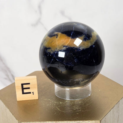 A polished dark blue and gold jasper stone sphere displayed on a clear acrylic stand atop a light wood pedestal, with a small Scrabble tile showing the letter E next to it against a white marble background.