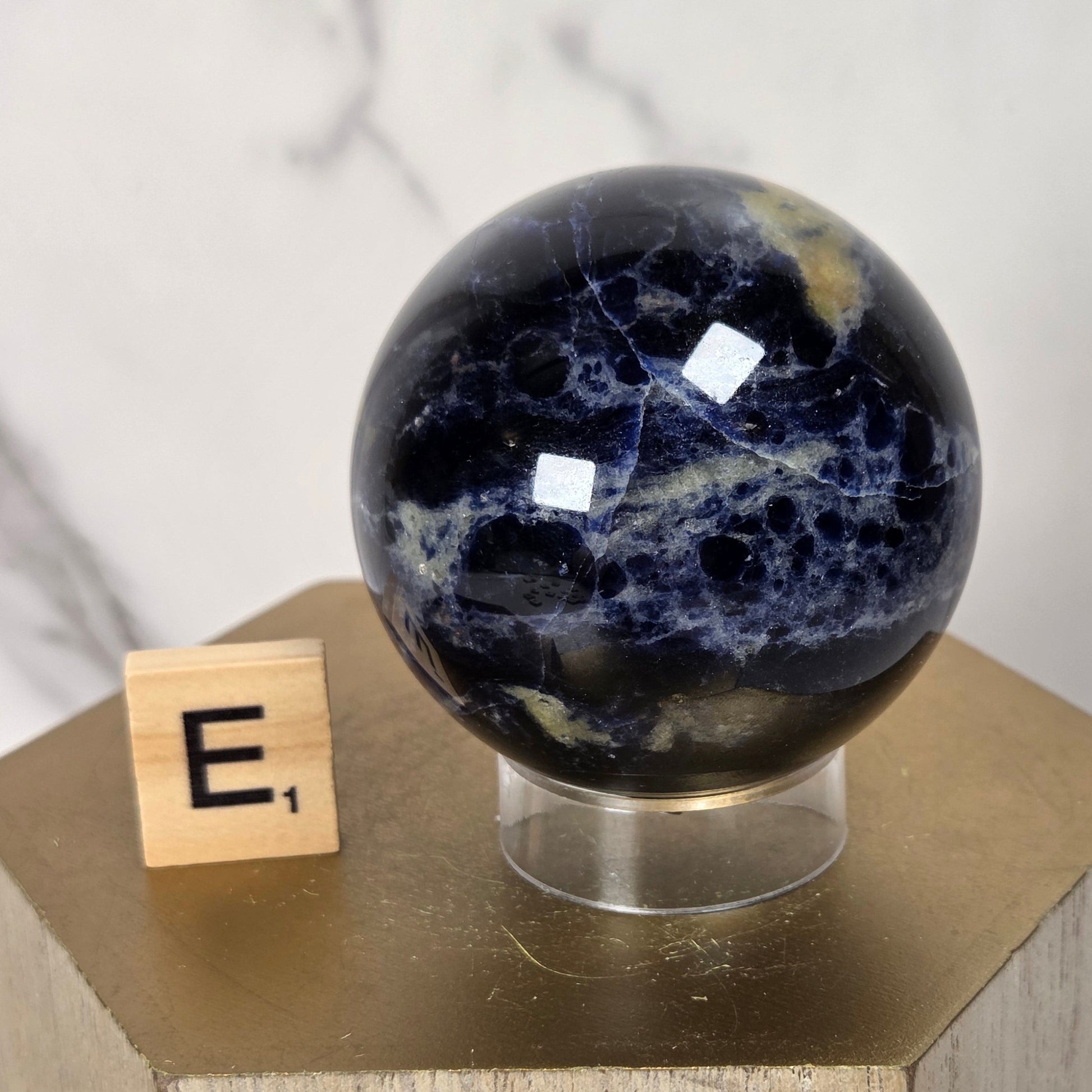 A polished dark blue and black stone sphere with white veining sits on a clear stand atop a gold-toned wooden block, with a small Scrabble tile showing the letter E next to it against a light marble background.