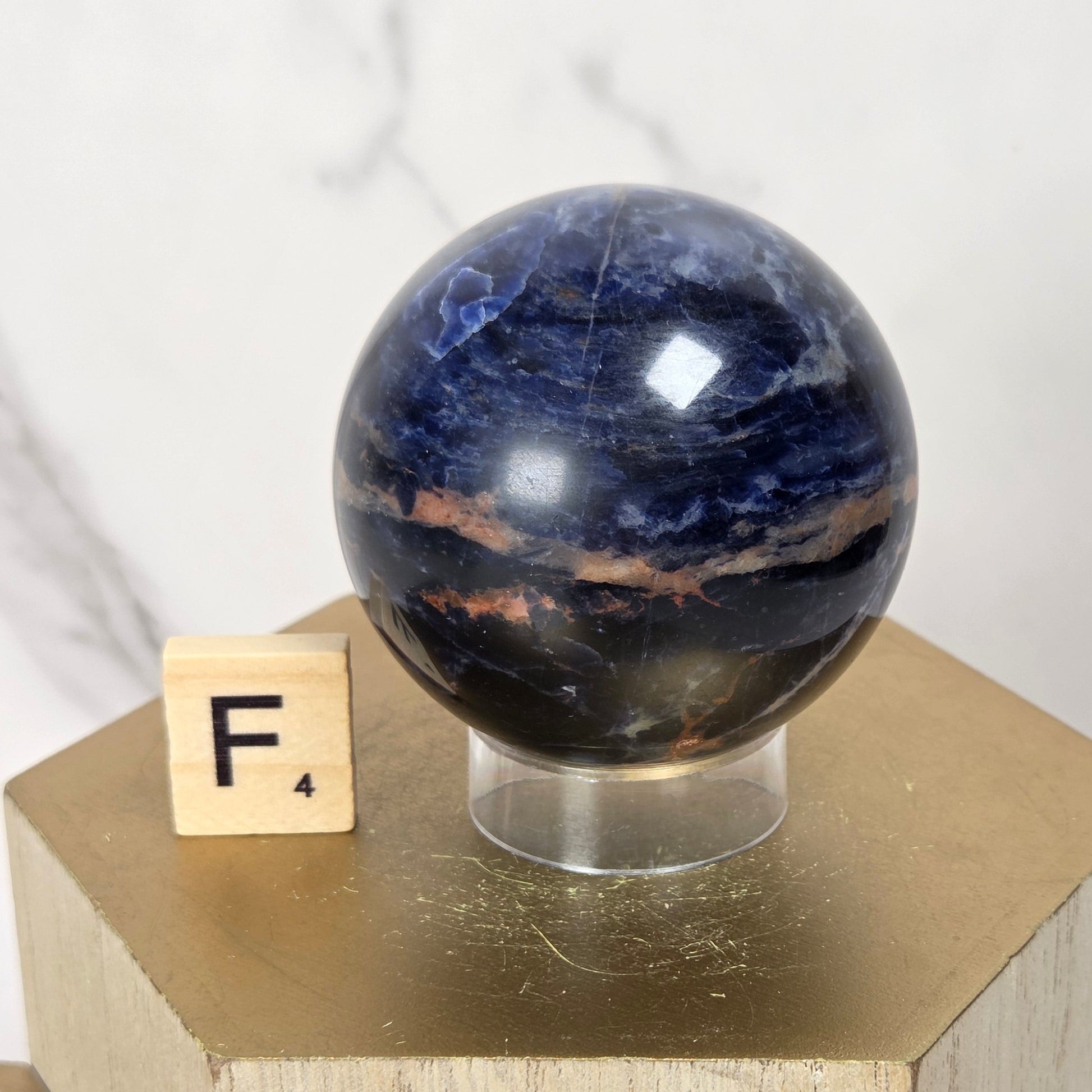 A polished dark blue and black stone sphere with coppery bands, displayed on a clear stand atop a gold geometric pedestal, with a small Scrabble tile showing 'F4' beside it.