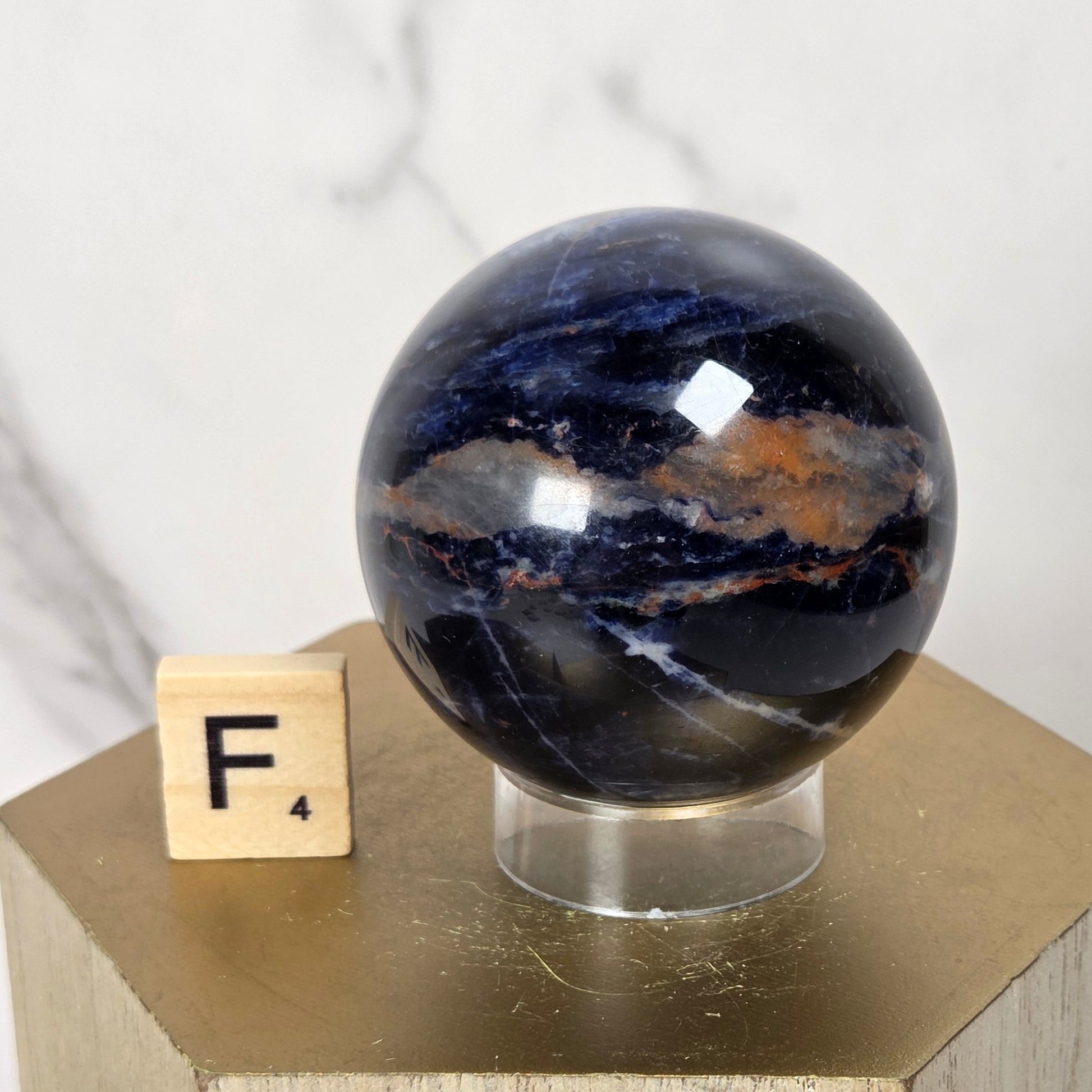 A polished dark blue and black stone sphere with natural banding, displayed on a clear stand atop a gold geometric pedestal next to a small wooden letter block.