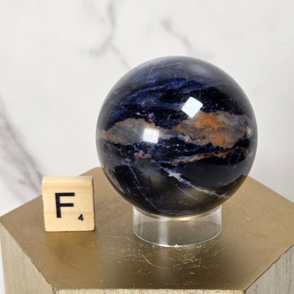 A polished dark blue and black stone sphere with natural banding, displayed on a clear stand atop a gold geometric pedestal next to a small wooden letter block.