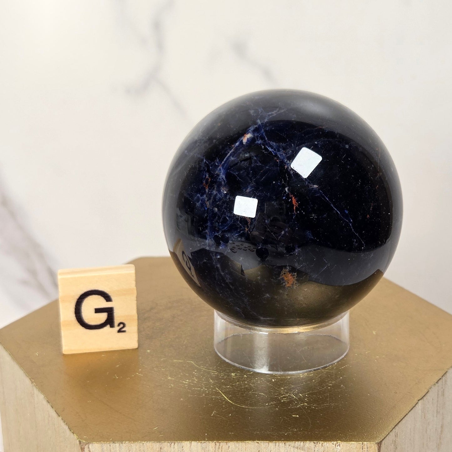A polished dark blue lapis lazuli sphere on a clear acrylic stand, displayed next to a small wooden Scrabble tile on a gold hexagonal pedestal against a white marble background.
