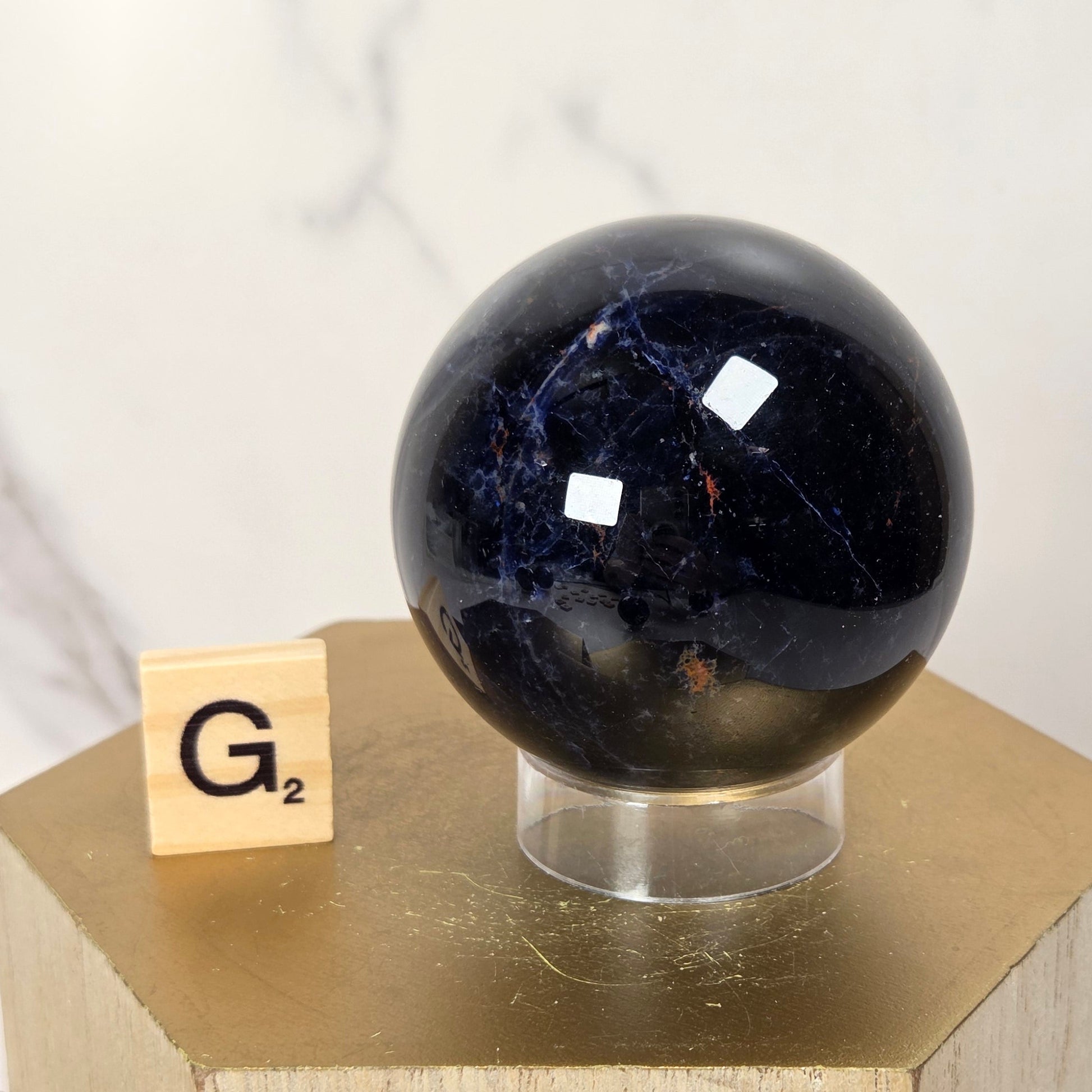 A polished dark blue lapis lazuli sphere on a clear acrylic stand, displayed next to a small wooden Scrabble tile on a gold hexagonal pedestal against a white marble background.