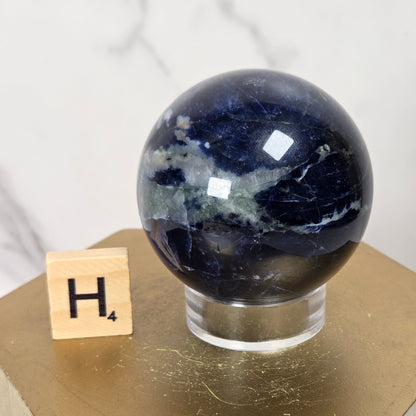 A polished dark blue selenite sphere with white and gray natural banding, displayed on a clear acrylic stand atop a gold hexagonal pedestal next to a small wooden letter tile.