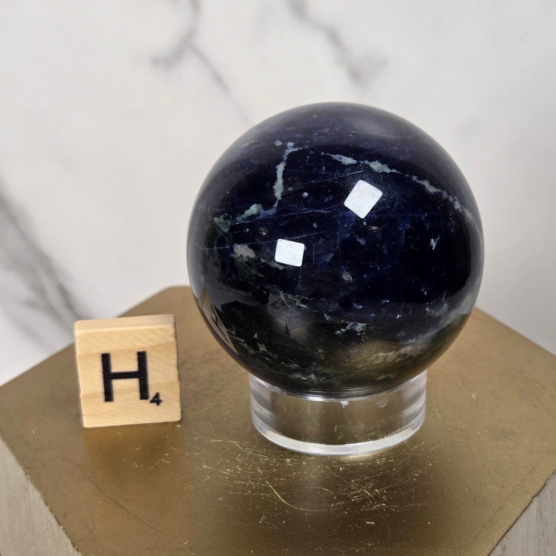 A polished dark blue selenite crystal sphere with white veining, displayed on a clear acrylic stand atop a gold hexagonal pedestal next to a wooden Scrabble tile.