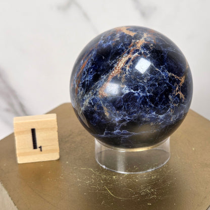 A polished dark blue and black stone sphere with natural gold veining, displayed on a clear acrylic stand atop a hexagonal gold pedestal. A small wooden block with the number 1 is next to it, set against a light marble-patterned background.