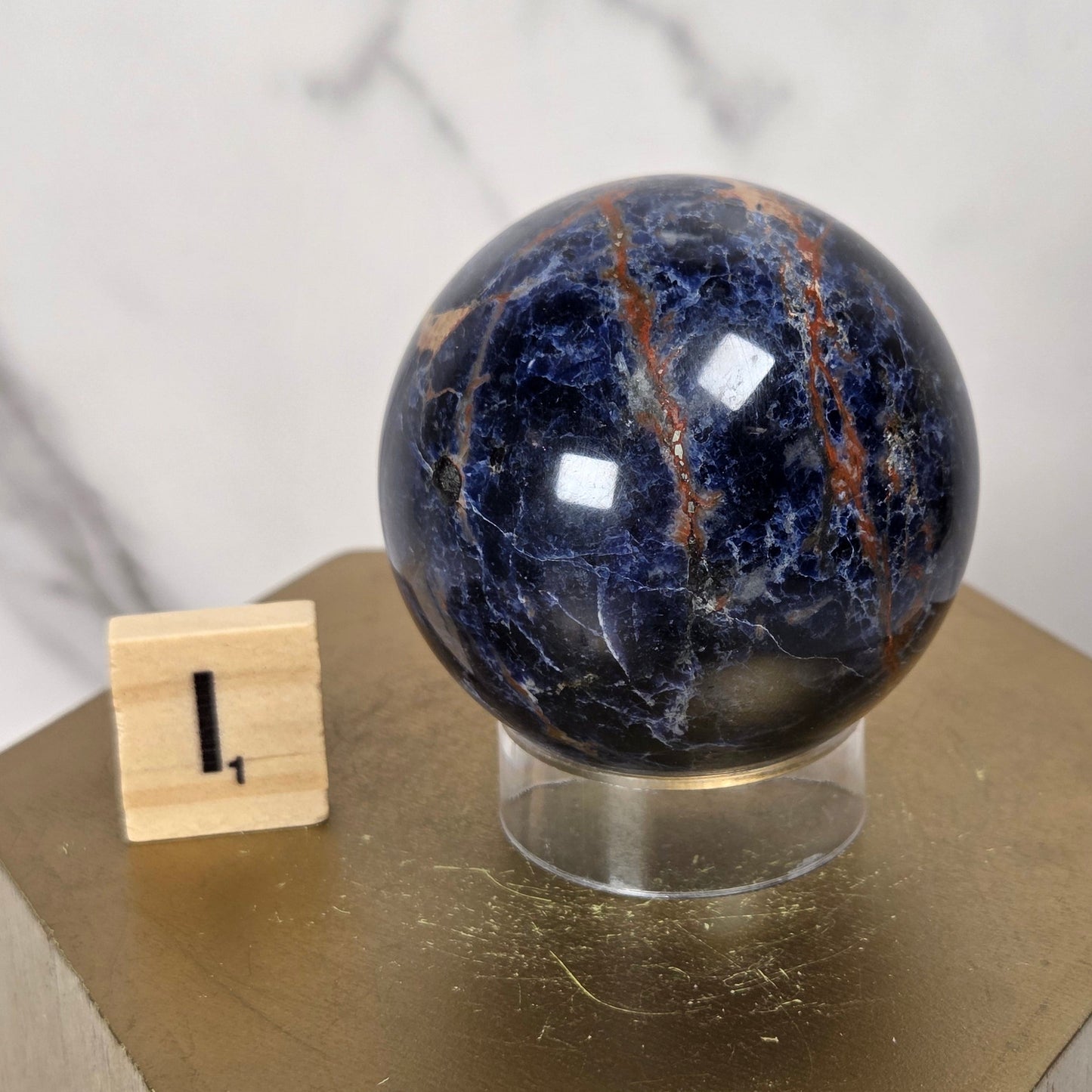 A polished dark blue selenite sphere with orange and white veining, displayed on a clear acrylic stand atop a gold hexagonal pedestal, with a small wooden block showing the number 1.