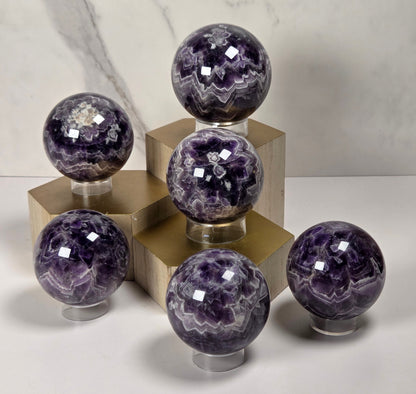 Five polished purple amethyst crystal spheres displayed on small clear stands atop gold-colored geometric pedestals against a light marble-patterned background.
