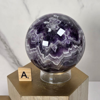 A polished purple amethyst crystal sphere with white and gray banding, displayed on a clear stand atop a gold hexagonal pedestal. A small wooden letter tile with the letter 'A' sits beside it against a light marble-patterned background.