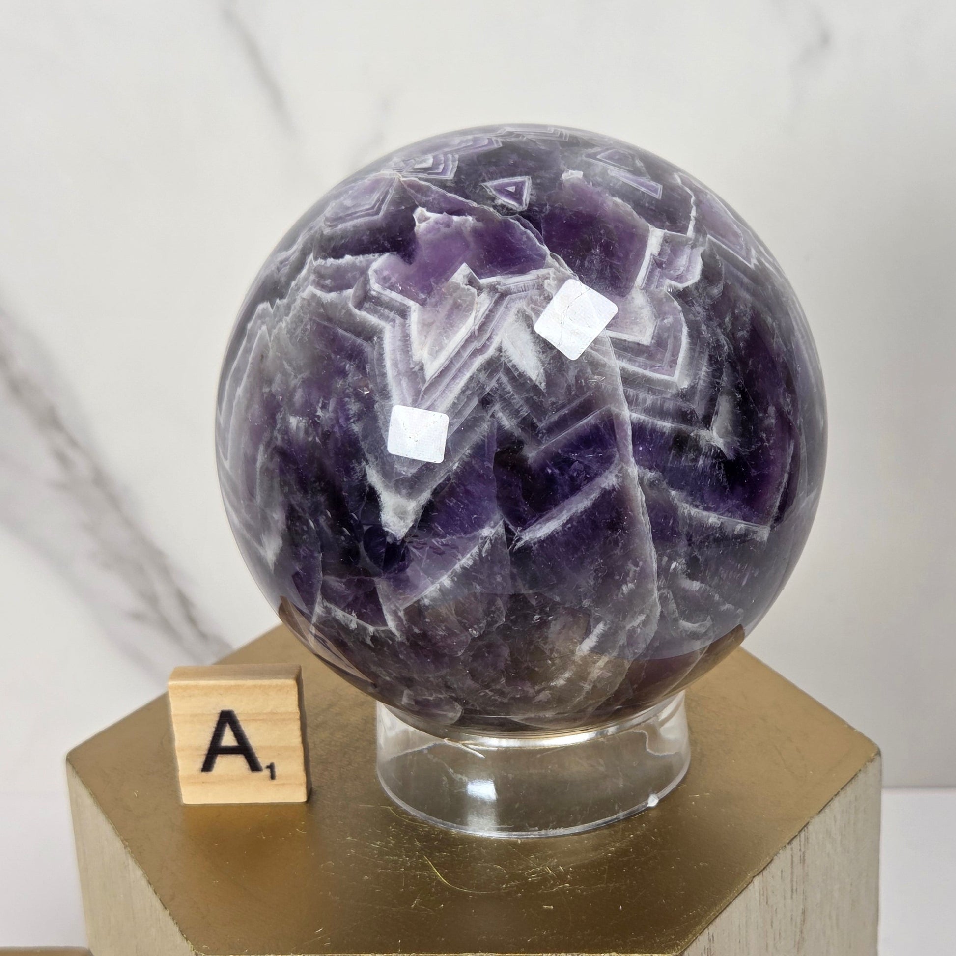 A polished purple amethyst crystal sphere with white and darker purple banding, displayed on a clear glass stand atop a gold hexagonal pedestal. A small wooden letter tile with the letter 'A' sits beside the base.