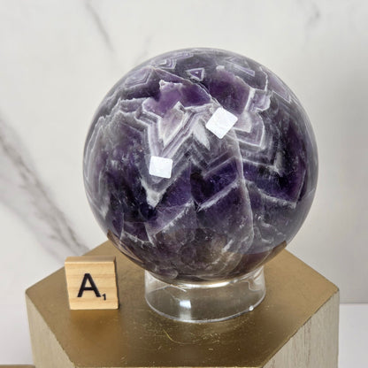 A polished purple amethyst crystal sphere with white and darker purple banding, displayed on a clear glass stand atop a gold hexagonal pedestal. A small wooden letter tile with the letter 'A' sits beside the base.