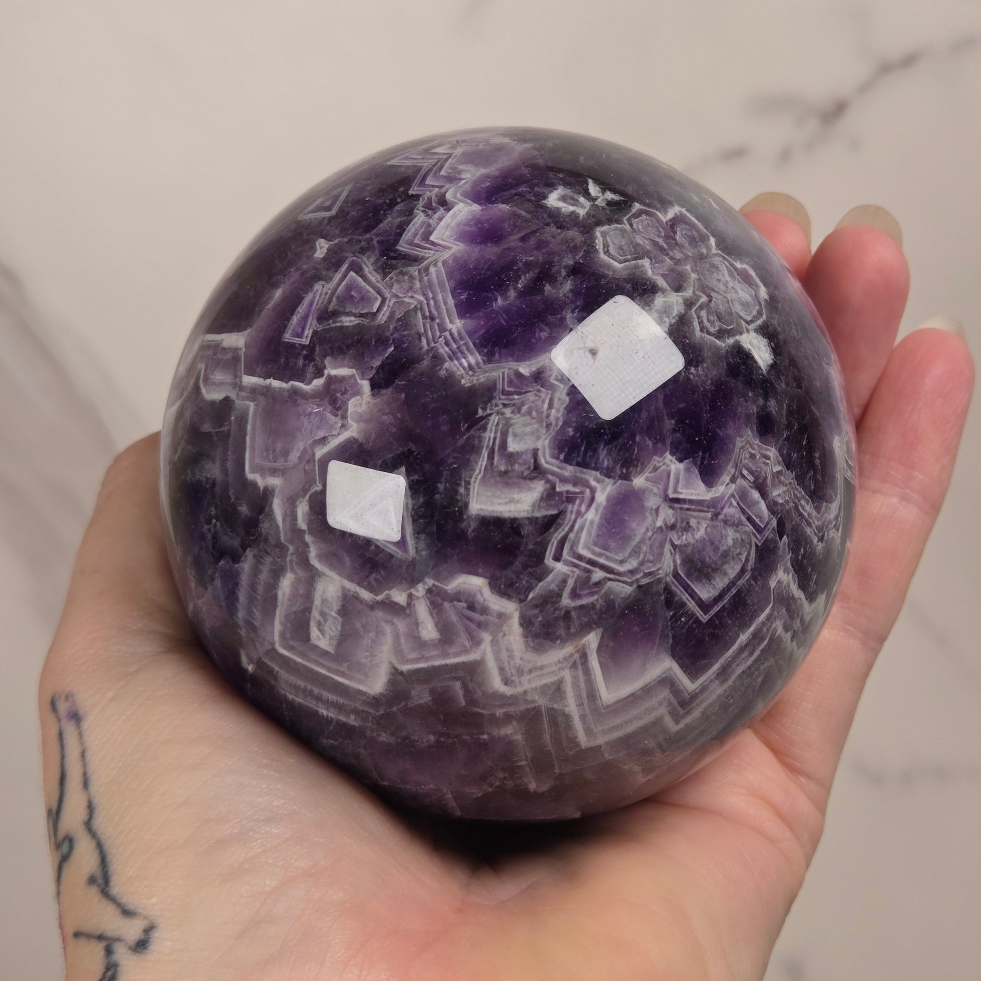 A hand holding a polished purple amethyst crystal sphere with natural geometric patterns, set against a light marble background.