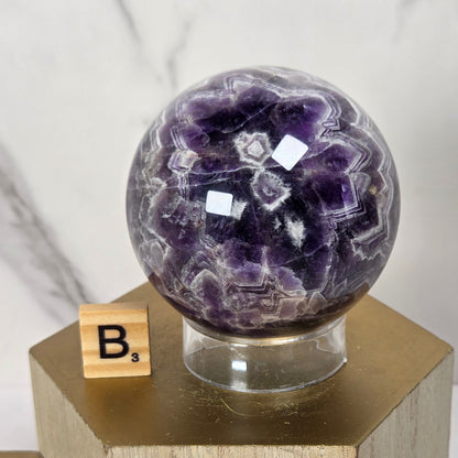A polished purple amethyst geode sphere displayed on a clear glass stand atop a gold hexagonal pedestal, with a wooden Scrabble tile showing 'B' beside it against a light marble background.
