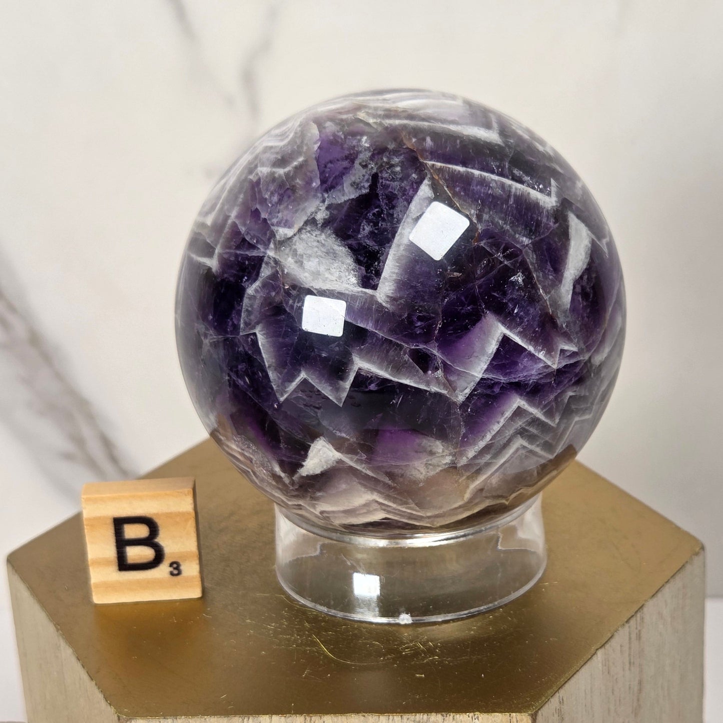 A faceted amethyst crystal sphere on a clear glass stand, displayed on a gold hexagonal pedestal with a small Scrabble tile showing the letter B.