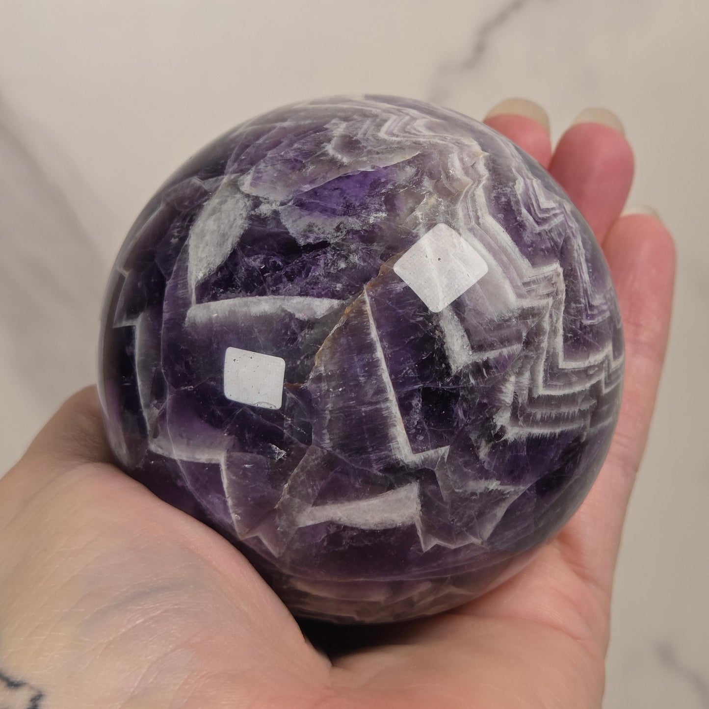 A hand holding a polished purple and white agate crystal sphere against a light marble background.