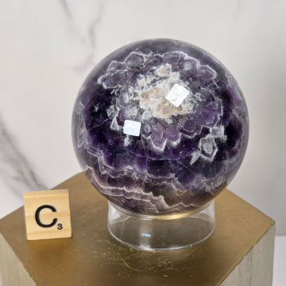 A polished purple amethyst geode sphere displayed on a clear acrylic stand atop a gold hexagonal pedestal, with a small wooden Scrabble tile showing the letter C in front, set against a light marble-patterned background.