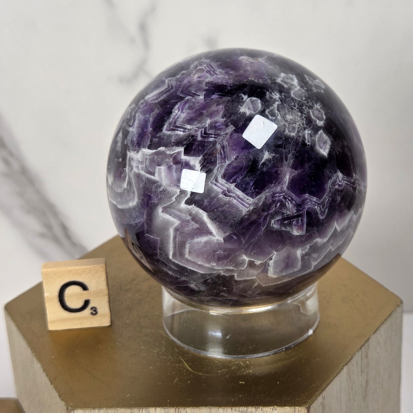 A polished purple amethyst crystal sphere on a clear acrylic stand, displayed on a hexagonal wooden pedestal with a small Scrabble tile showing the letter C.