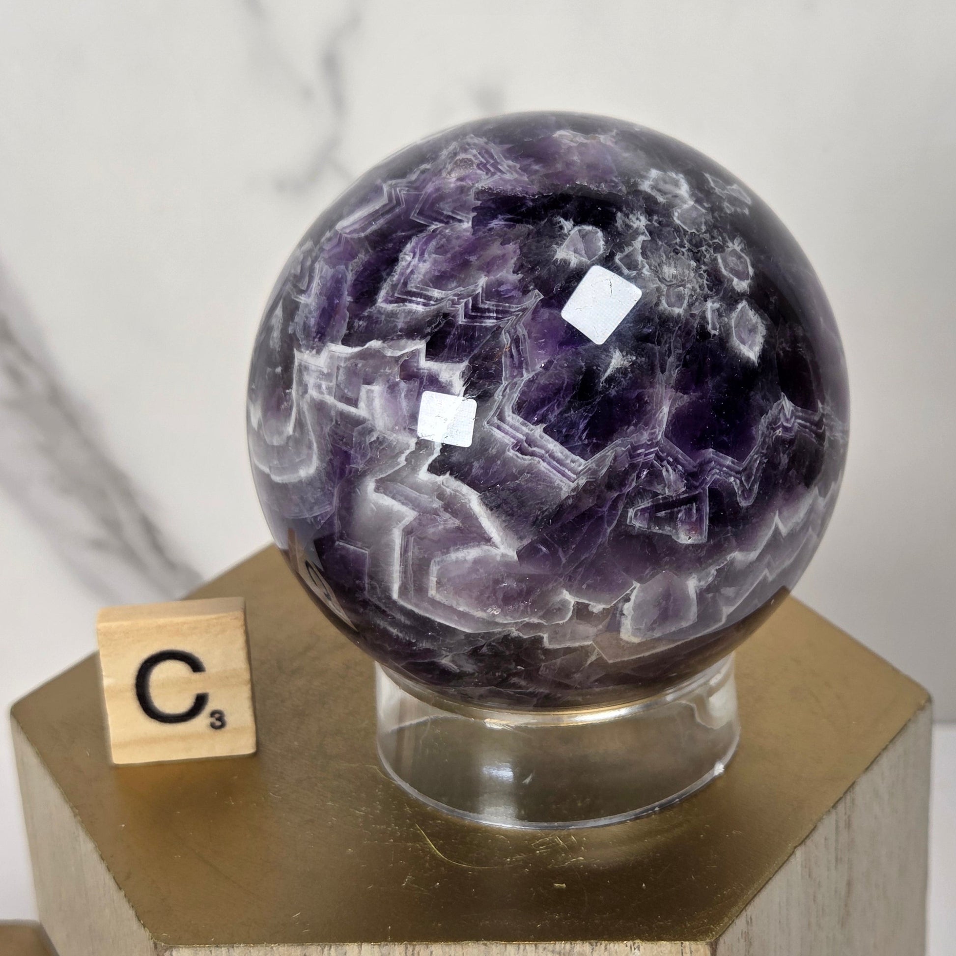 A polished purple amethyst crystal sphere on a clear acrylic stand, displayed on a hexagonal wooden pedestal with a small Scrabble tile showing the letter C.