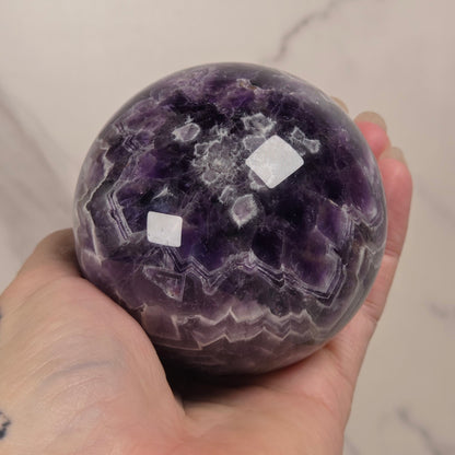 A hand holding a polished purple amethyst geode sphere with visible white quartz crystal formations, set against a light marble-patterned background.
