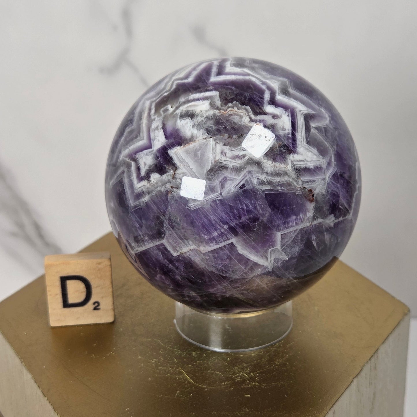 A polished purple amethyst crystal sphere with white geometric banding, displayed on a clear stand atop a gold hexagonal pedestal. A small wooden Scrabble tile showing 'D2' sits beside it against a light marble-patterned background.