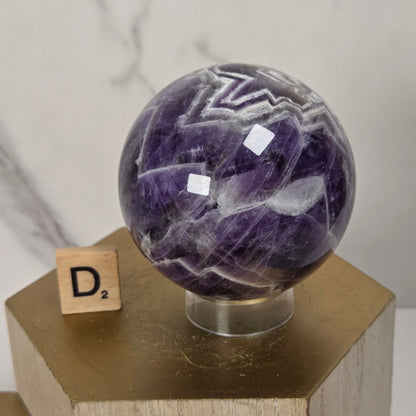 A polished purple amethyst crystal sphere with white agate banding, displayed on a clear stand atop a gold hexagonal pedestal next to a wooden Scrabble tile.