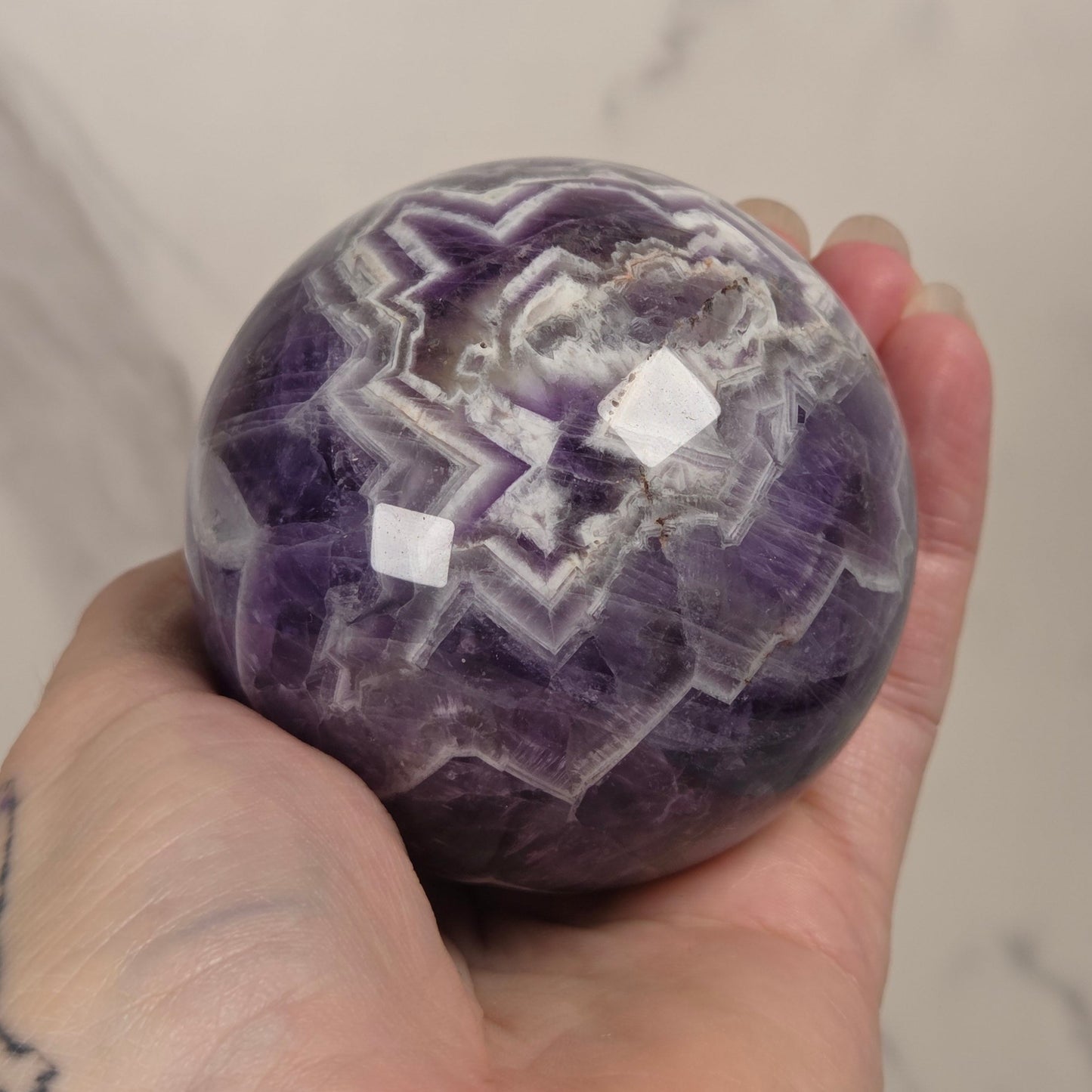 A hand holding a polished purple and white agate sphere against a light marble background.