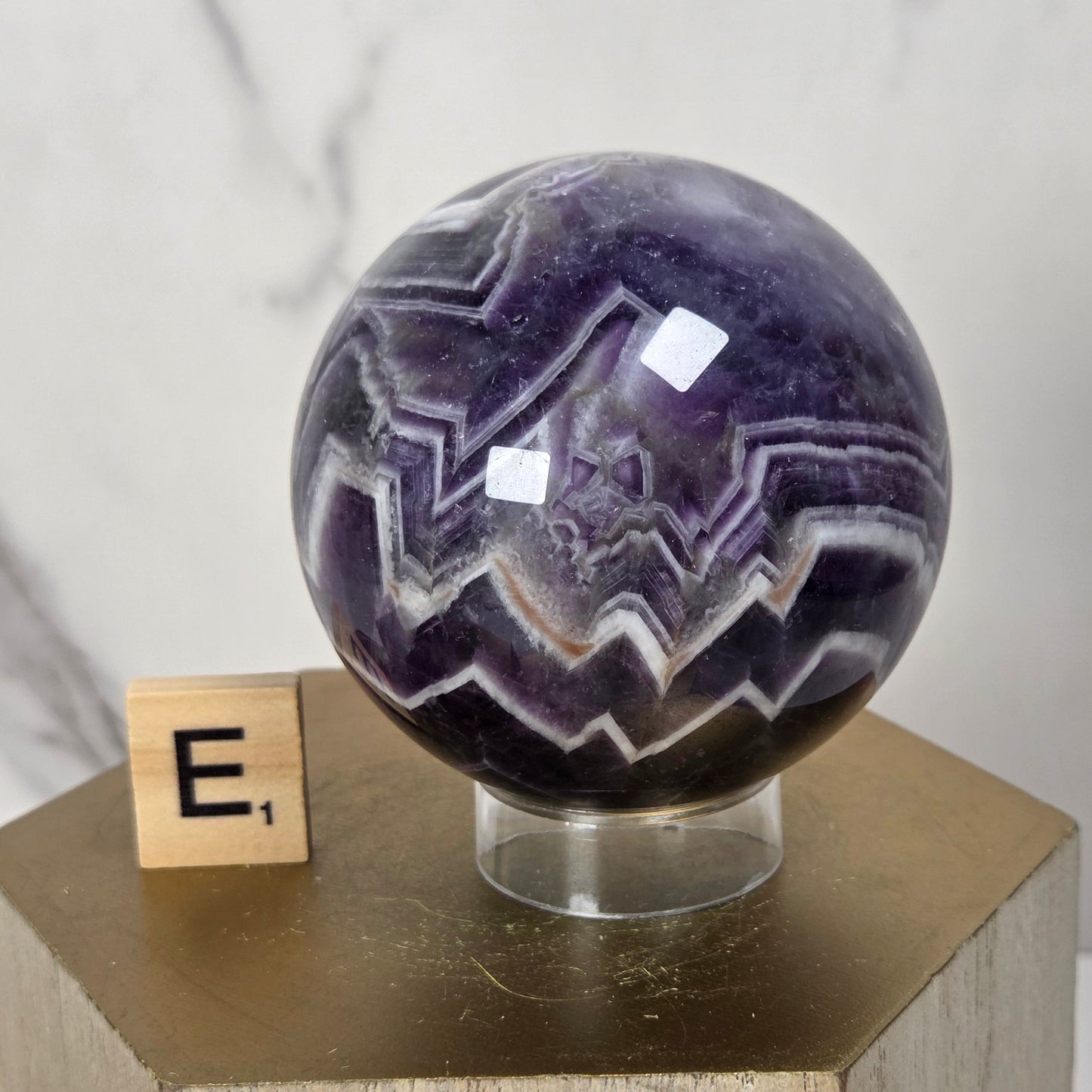 A polished purple amethyst crystal sphere with white and dark purple jagged banding, displayed on a clear stand atop a wooden pedestal next to a small wooden block with the letter E.