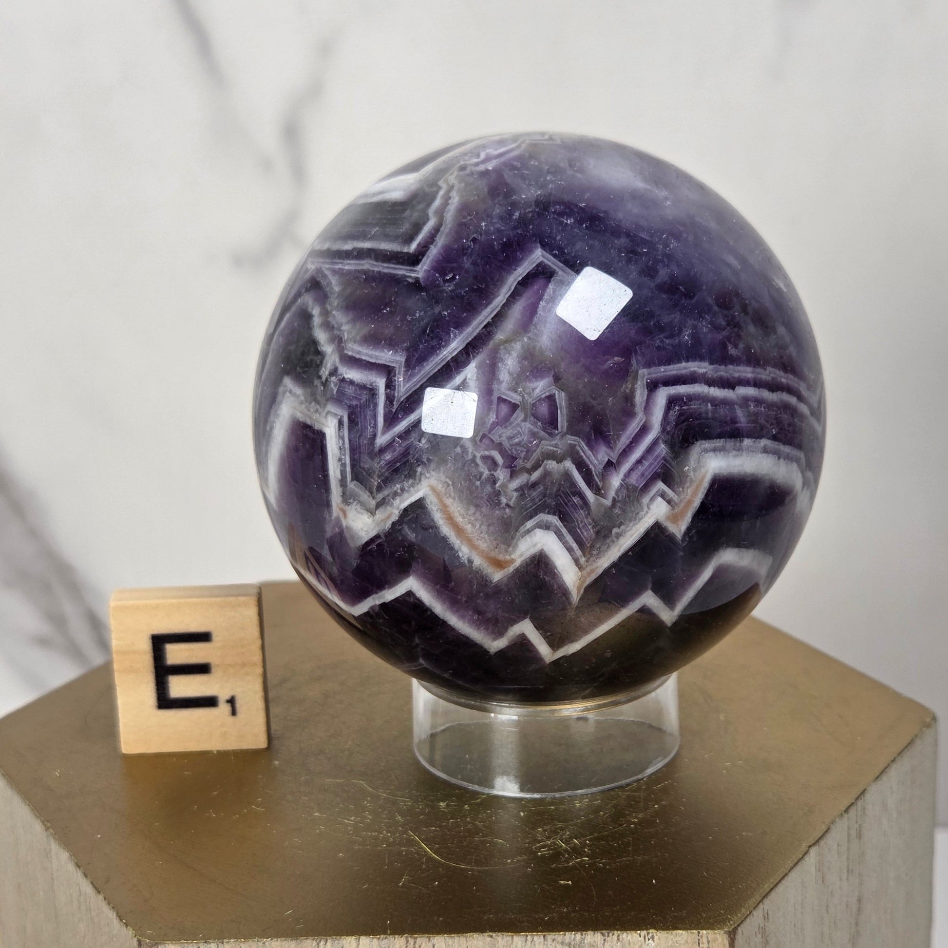 A polished purple amethyst crystal sphere with white and dark purple jagged banding, displayed on a clear stand atop a wooden pedestal next to a small wooden block with the letter E.