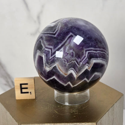 A polished purple amethyst crystal sphere with white and dark purple jagged banding, displayed on a clear stand atop a wooden pedestal next to a small wooden block with the letter E.