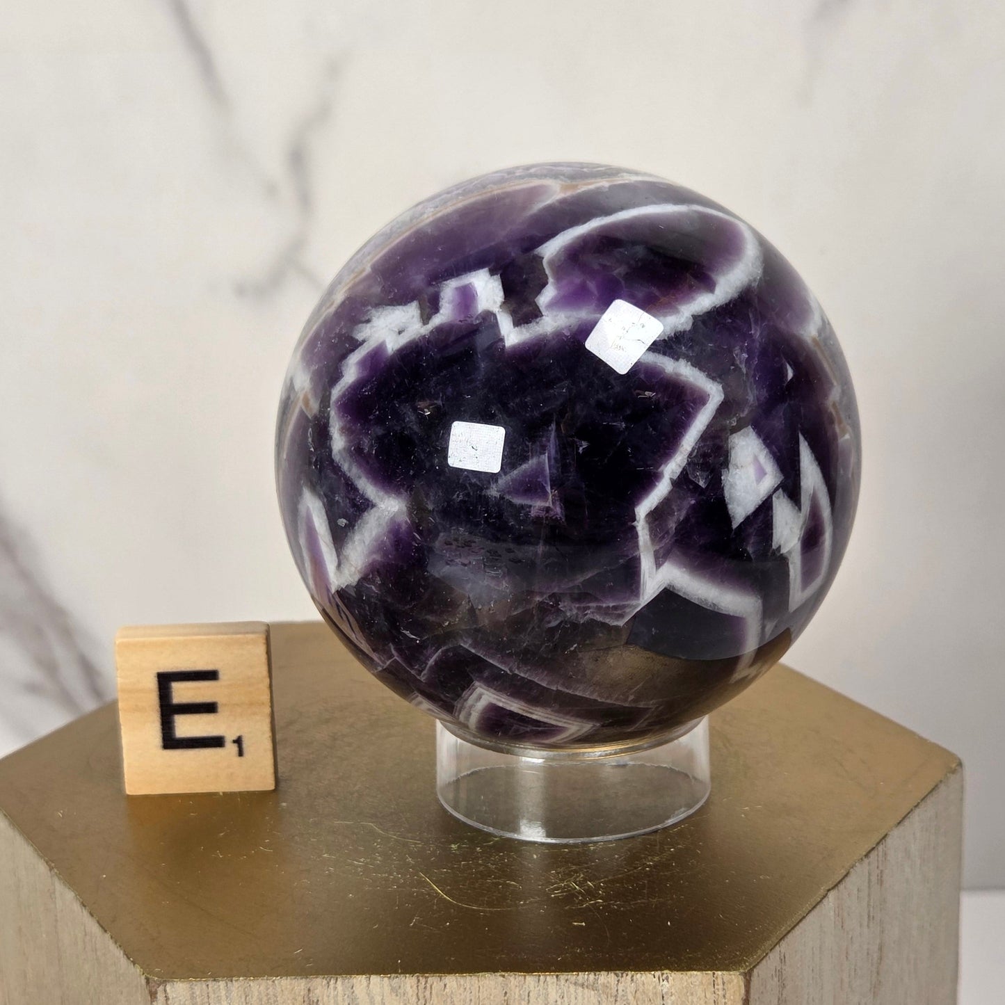 A polished purple and white amethyst crystal sphere displayed on a clear stand atop a wooden pedestal, with a small letter block showing 'E,' in the background against a light marble wall.