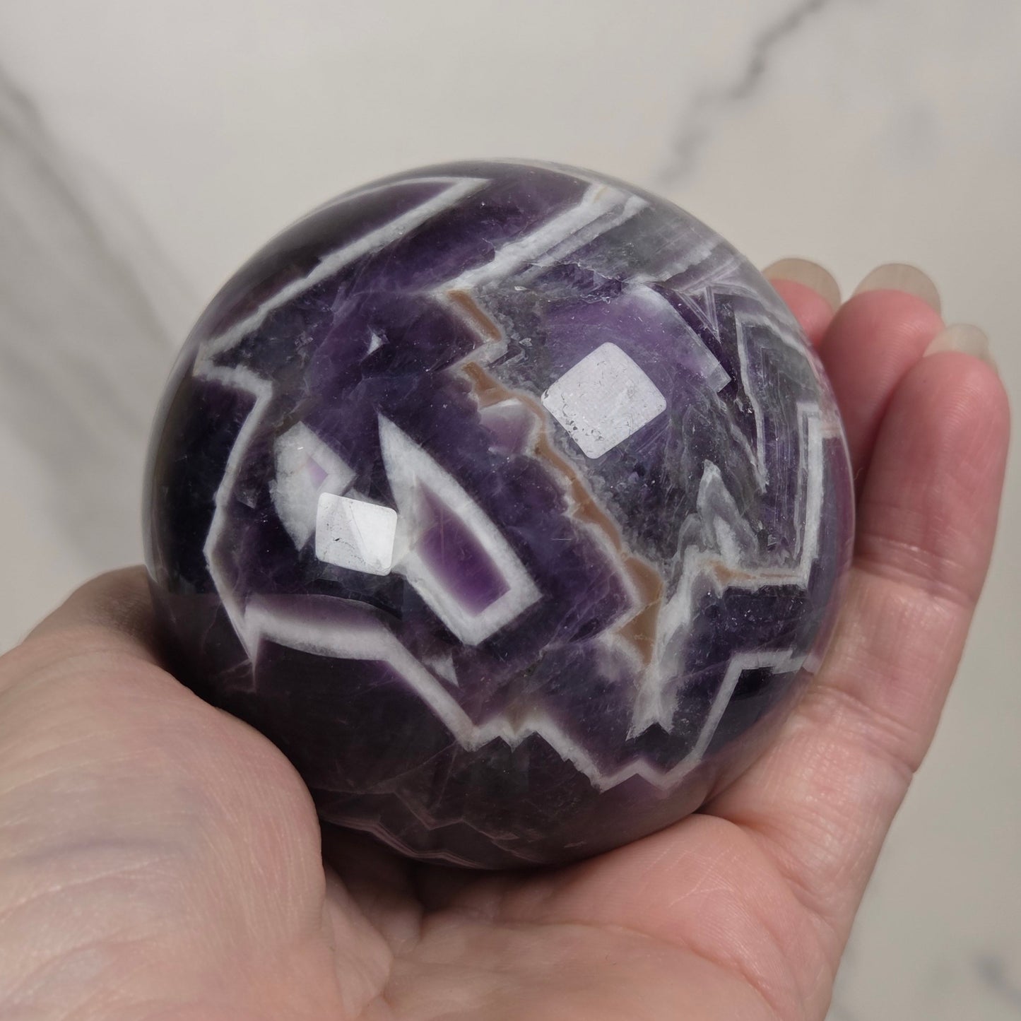 A hand holding a polished purple and white agate sphere against a light marble background.