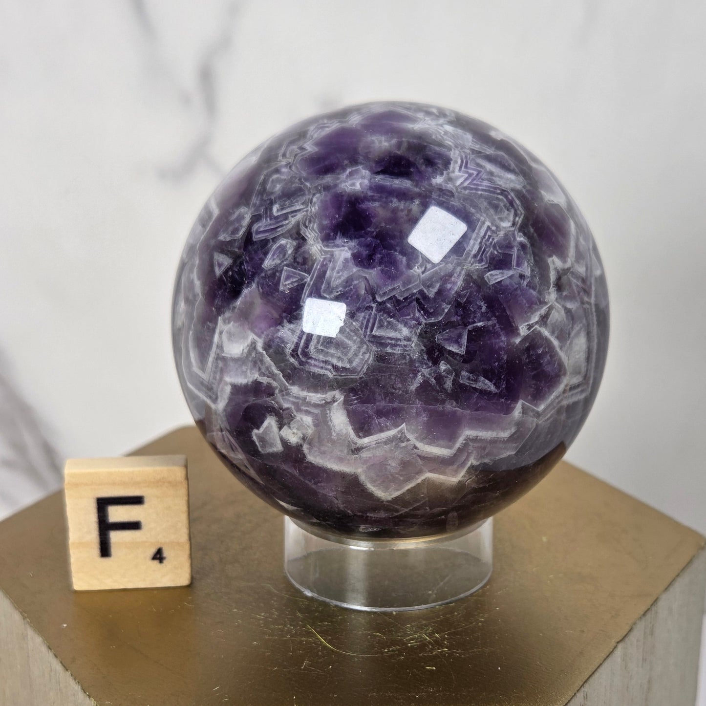 A polished purple amethyst crystal sphere on a clear acrylic stand, displayed on a gold hexagonal pedestal next to a small wooden letter block with the letter F.