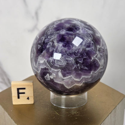 A polished purple amethyst crystal sphere on a clear acrylic stand, displayed on a gold hexagonal pedestal next to a small wooden letter block with the letter F.